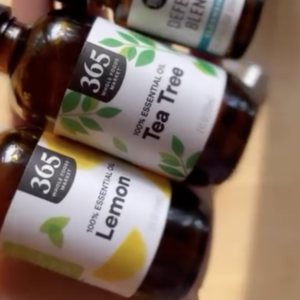 A person holds three brown bottles of essential oils. Labels read 365 Everyday Value Lemon, 365 Everyday Value Tea Tree, and Defend Blend. The background is blurred.